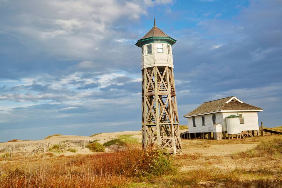 Un phare en bois, avec une maison blanche à proximité, entouré de dunes de sable et d’une végétation clairsemée.