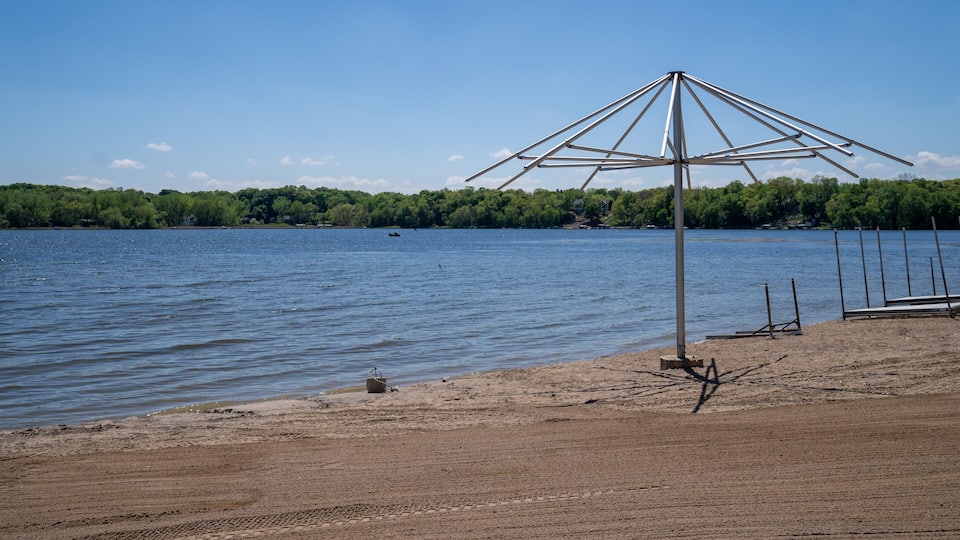 A beach with a metal umbrella structure, calm water, and a forested shoreline.