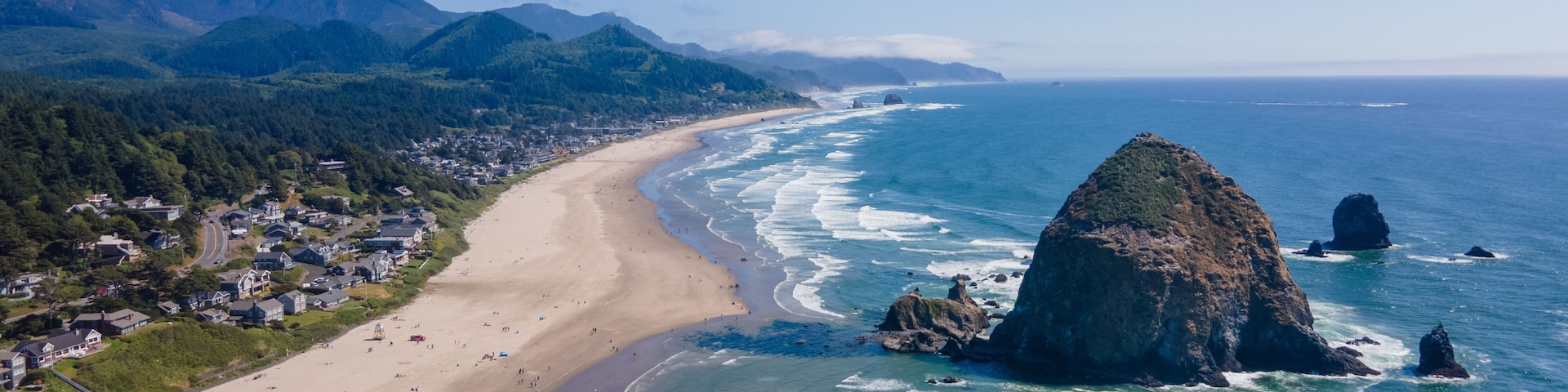 Uma vila costeira com praia, ondas do oceano e montanhas ao fundo.