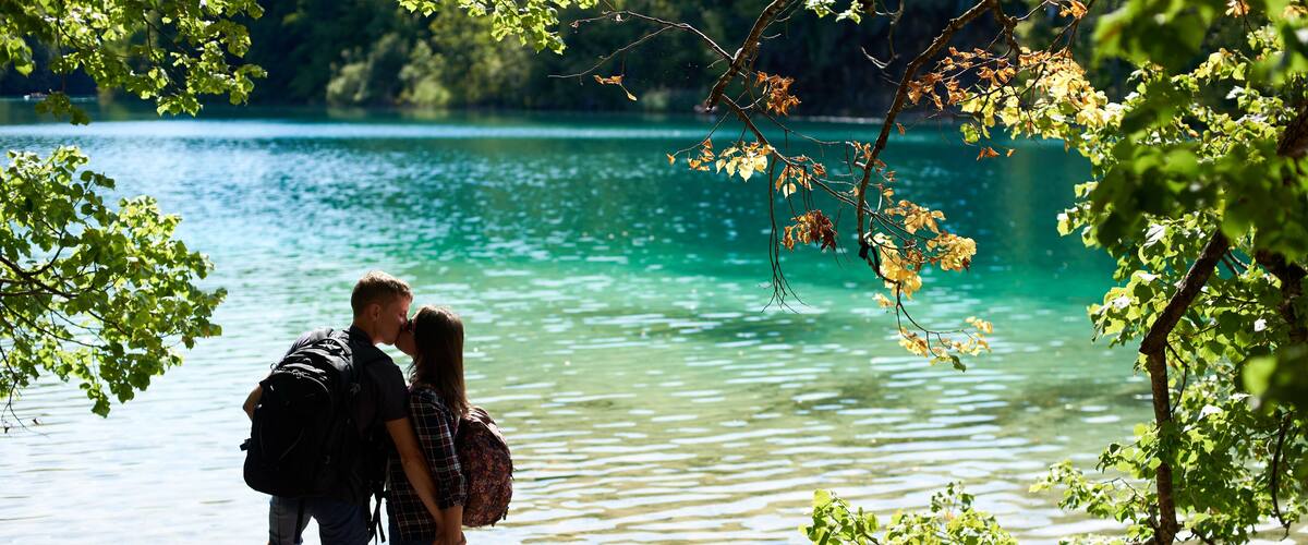 Back view of tourist young couple man and woman with backpacks standing on river bank, kissing each other, enjoying beautiful colorful spring panorama of lit by sun green forest and blue lake