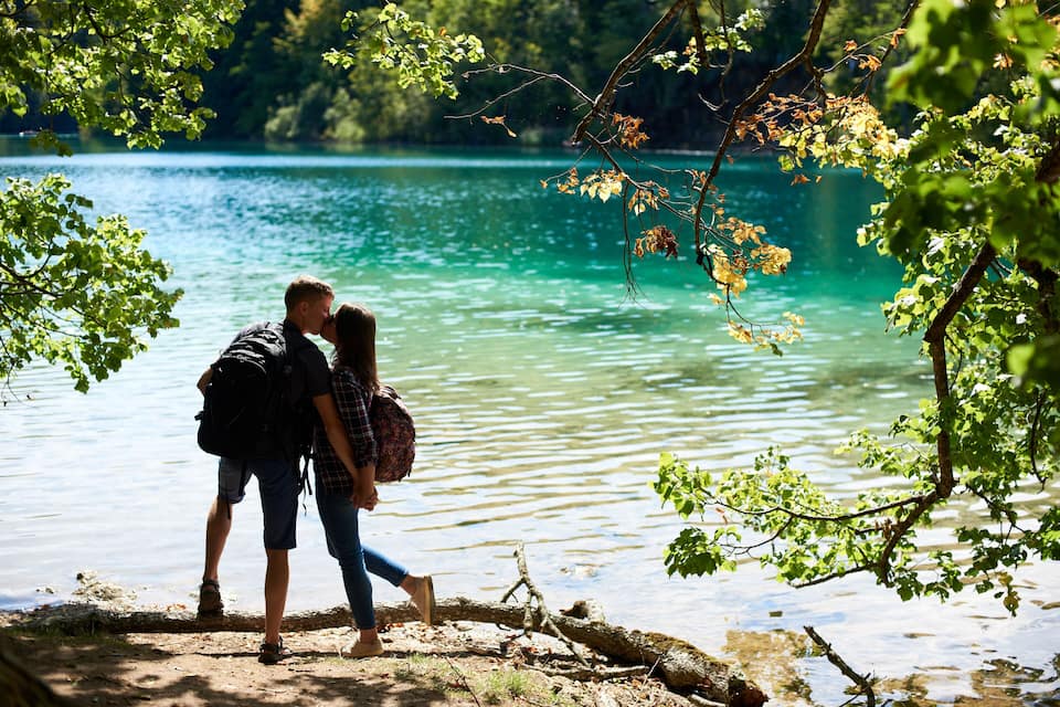 Un couple se tient près d’un lac dont l’eau est claire, entouré d’une végétation luxuriante.