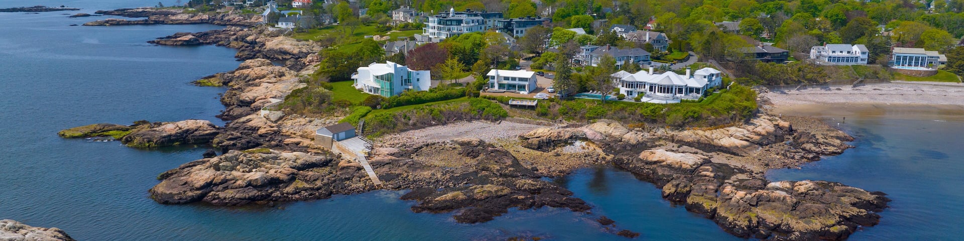 A coastal area with rocky shores, clear blue water, and several large houses surrounded by greenery.