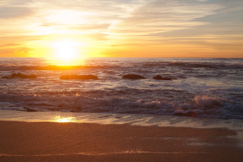 A beach at sunset with waves crashing onto the shore and the sun setting over the horizon.