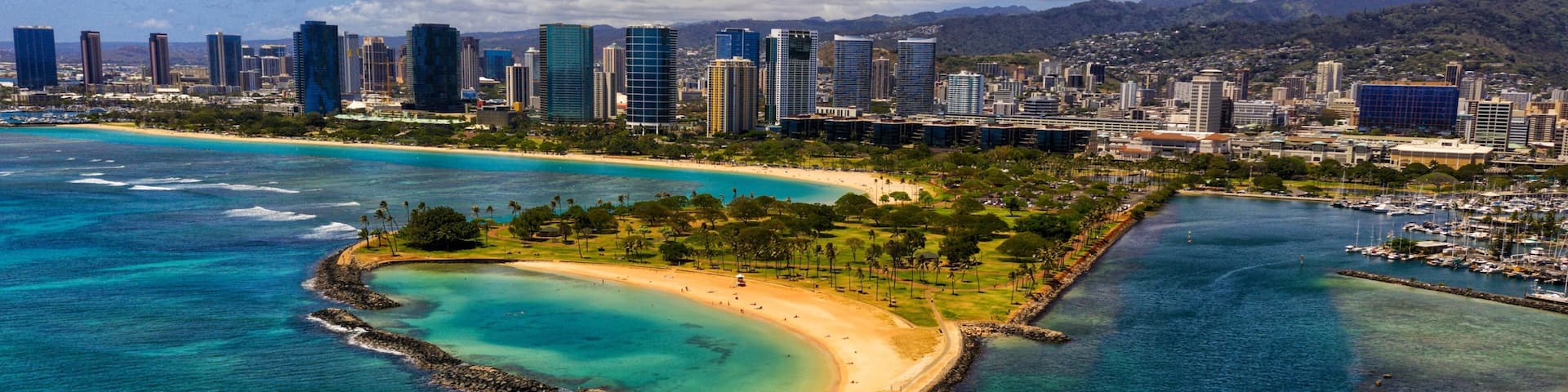 A coastal cityscape with a prominent beach, clear blue waters, and a backdrop of mountains.