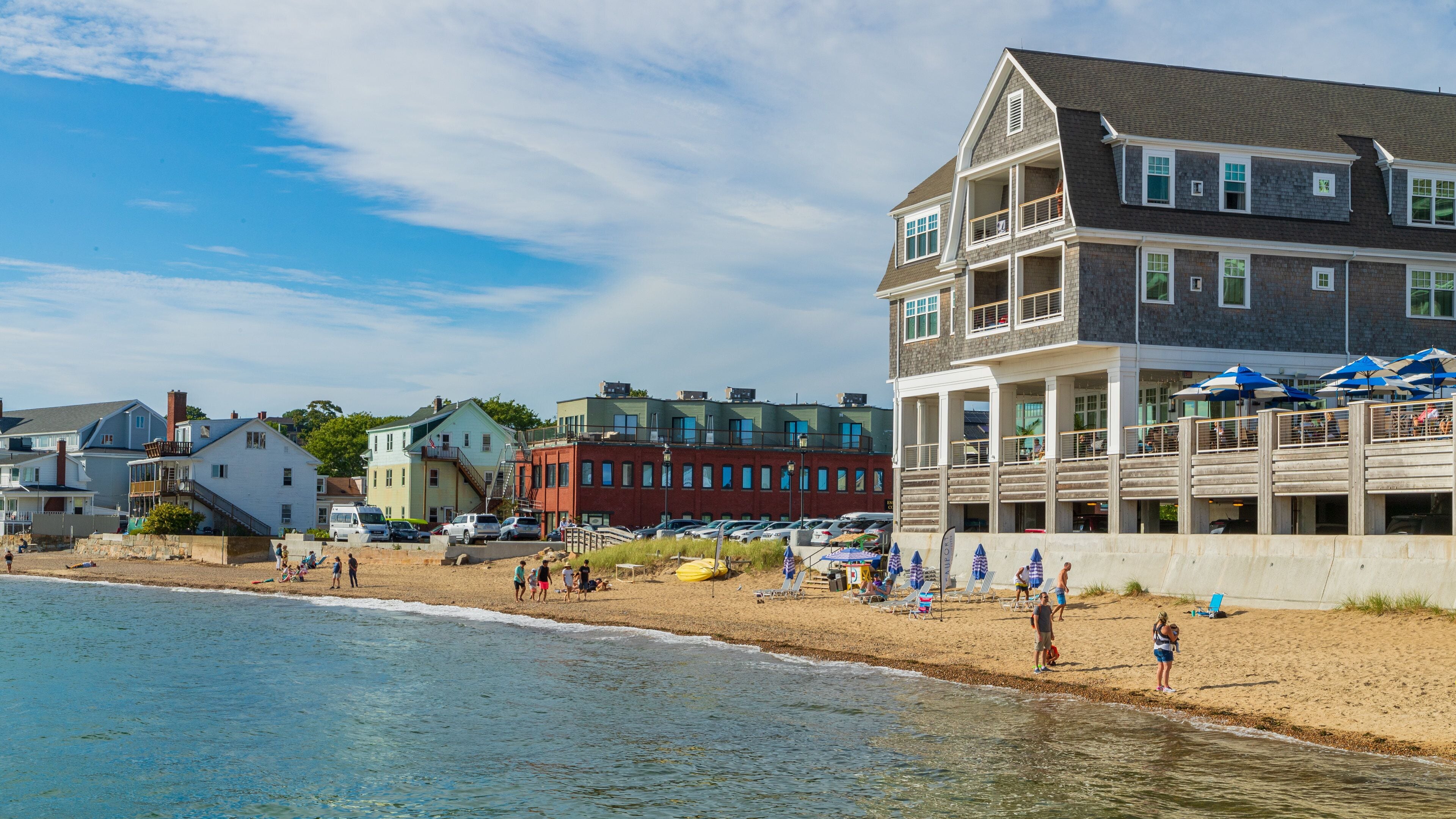 A beach with people, a wooden pier, and buildings in the background.
