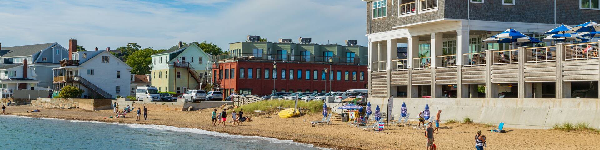 Una playa con gente, un muelle de madera y edificios al fondo.