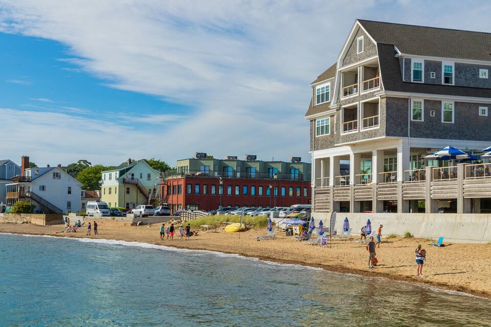 A beach with people, a wooden pier, and buildings in the background.