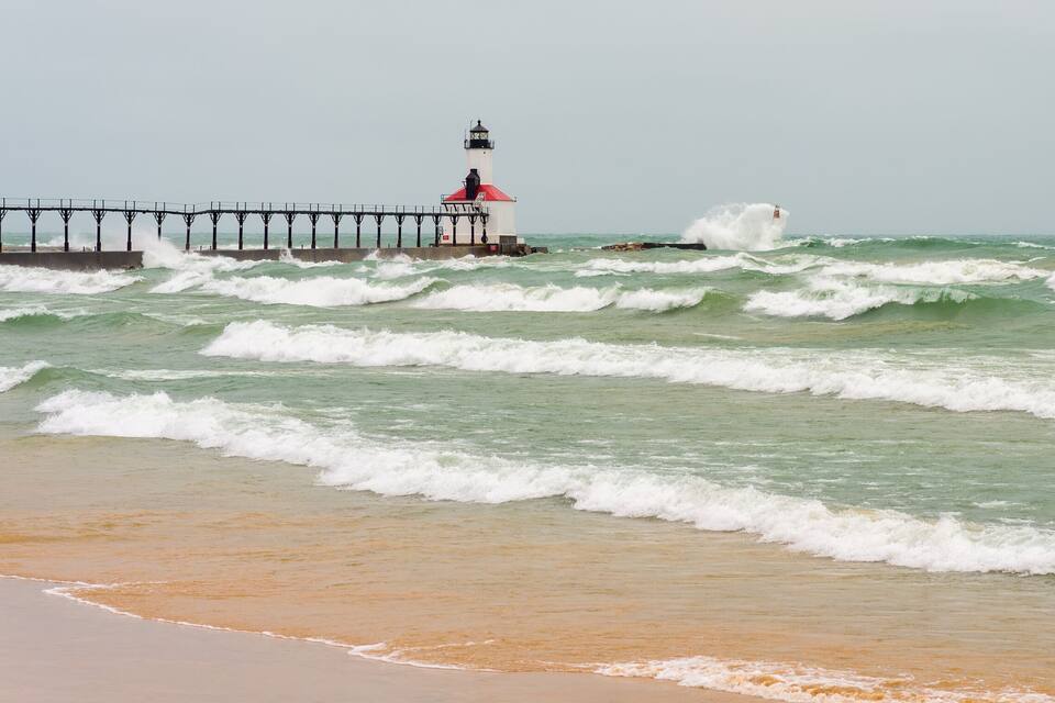 Una playa con olas rompiendo en la orilla, un muelle que se adentra en el agua y un faro al fondo.