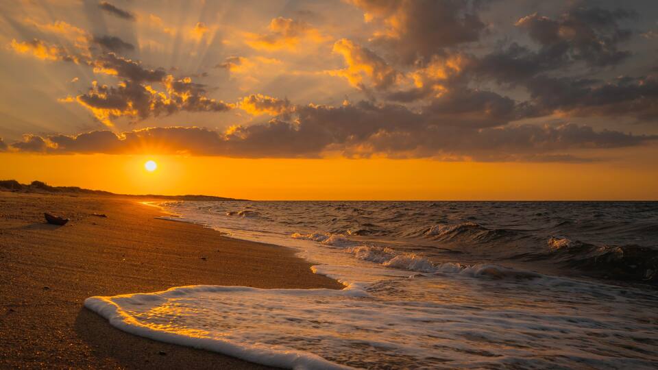 Una playa al atardecer con olas rompiendo en la orilla y el sol ocultándose en el horizonte.