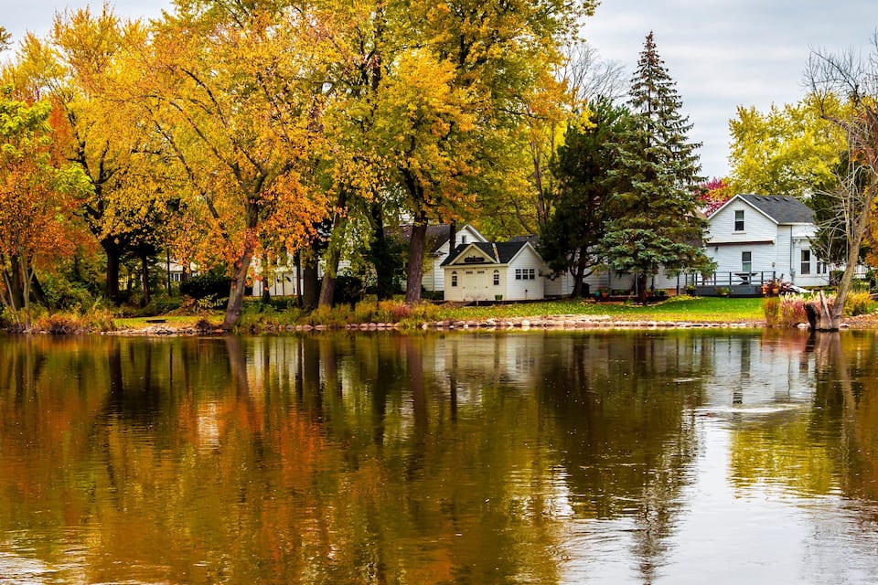 A lake with autumn-colored trees reflecting on the water, surrounded by houses.