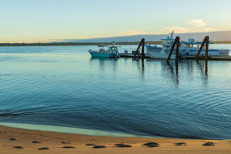 A calm harbor with several fishing boats docked, a sandy beach, and a distant shoreline with trees.