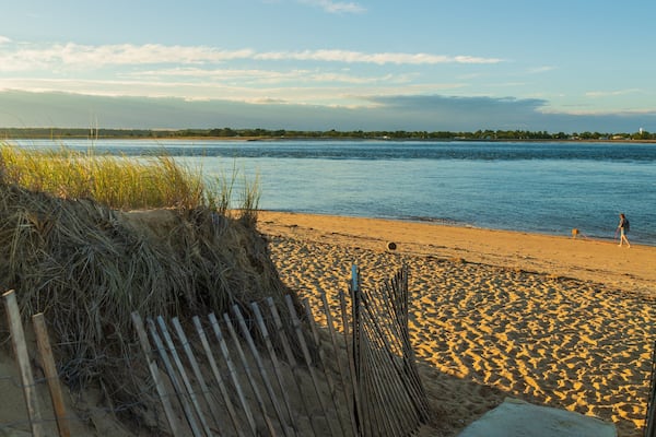 A sandy beach with a wooden fence and grass in the foreground, a calm river in the background, and a person walking a dog.