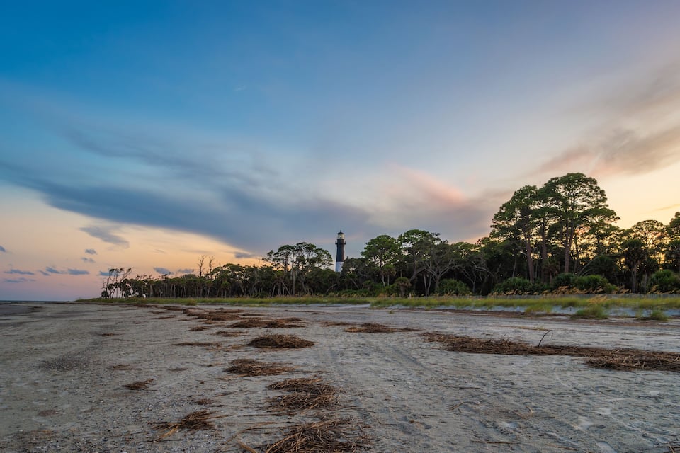 A beach with a lighthouse in the distance, surrounded by trees and a cloudy sky.