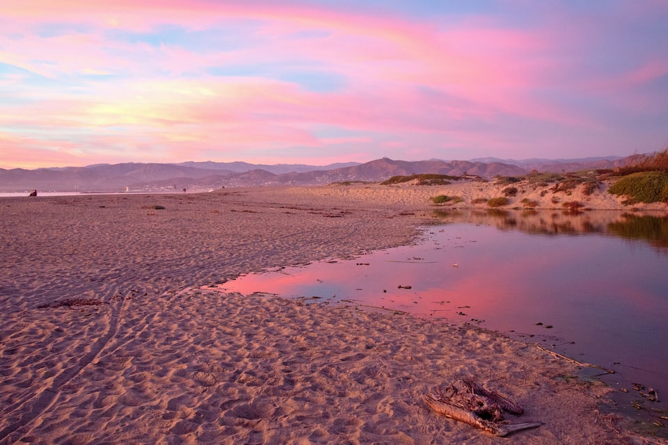 Ein Strand mit Dünen und ruhigem Wasser, das den Himmel spiegelt.