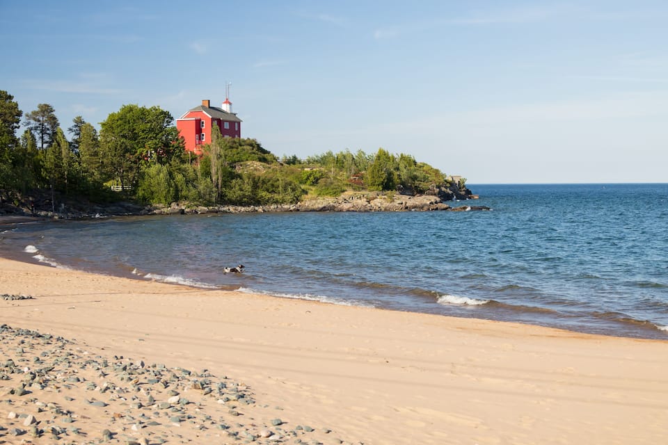 Una playa con una casa roja en una pequeña isla, rodeada de árboles y un litoral rocoso.