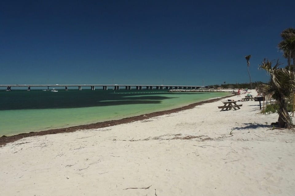 Een strand met helder turquoise water, een zandstrand en een lange pier die het water in loopt.
