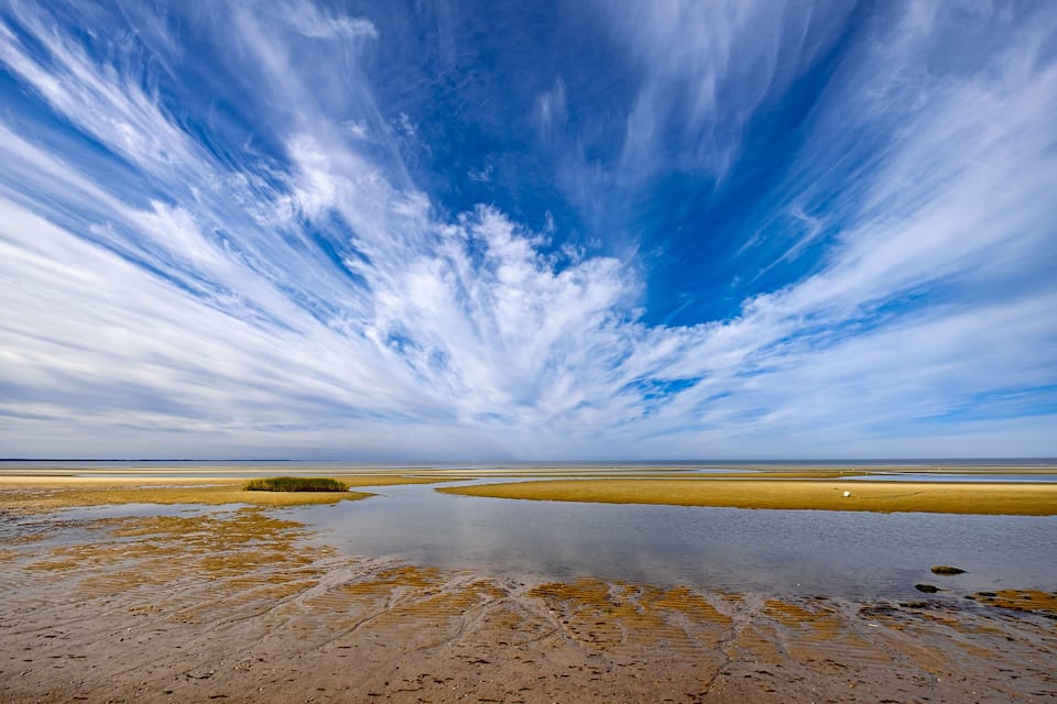 Paisatge costaner amb una àmplia platja de sorra, una petita massa d'aigua i un cel blau clar amb núvols.