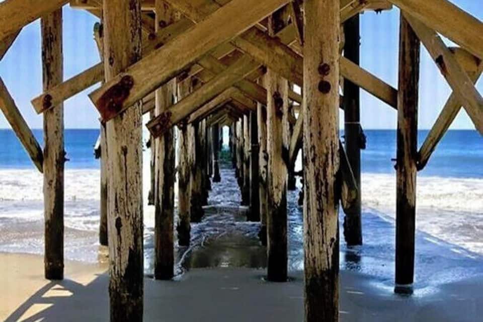 A wooden pier extending into the ocean with a clear view of the sandy beach and the sea.