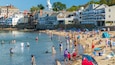 A crowded beach with people swimming and sunbathing, surrounded by colorful buildings and a church steeple.