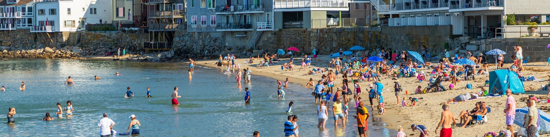 A crowded beach with people swimming and sunbathing, surrounded by colorful buildings and a church steeple.