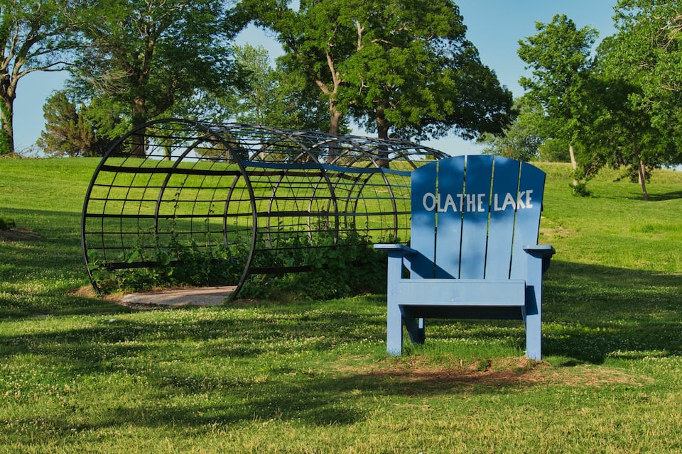 A blue Adirondack chair with 'OLATHE LAKE' written on it, set in a grassy park with trees and a metal fence in the background.