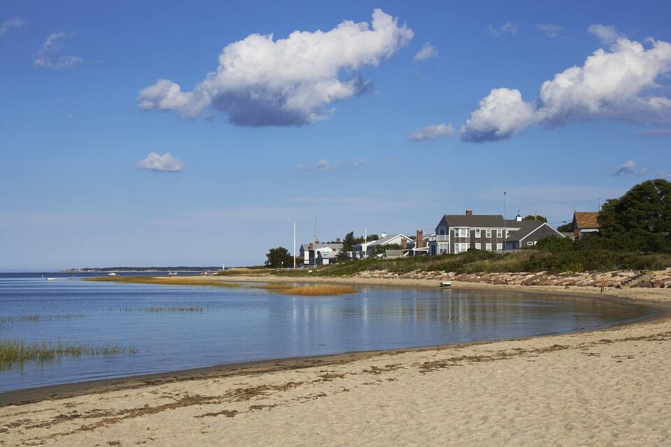 A beach with a calm sea, sandy shore, and houses in the background.