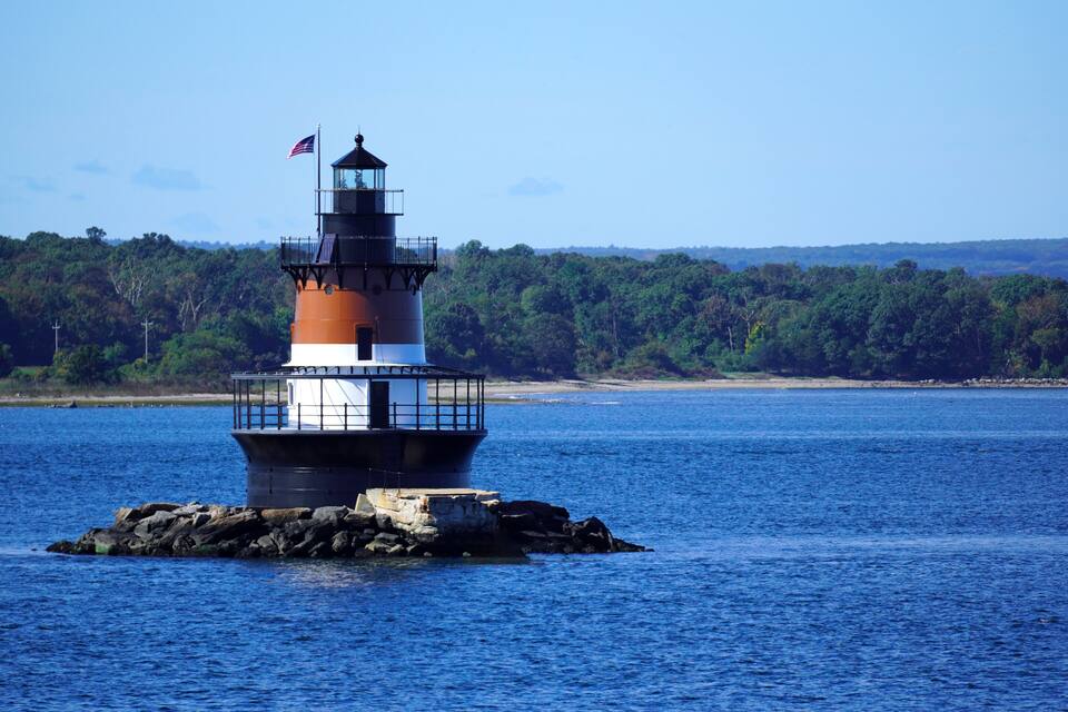 A lighthouse on a rocky outcrop in a body of water, surrounded by dense forest and a clear sky.