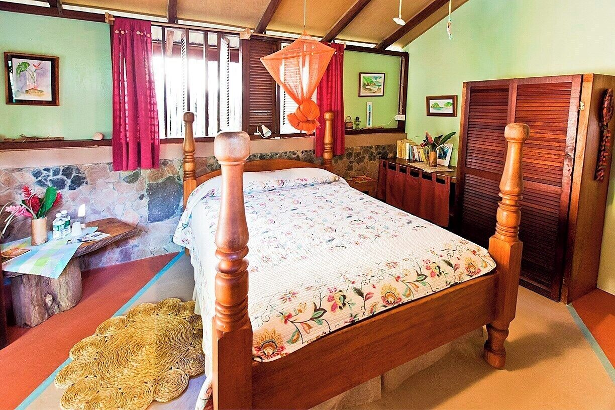 A wooden bed with a floral bedspread, a woven mat, a desk with a plant, and a wooden wardrobe.