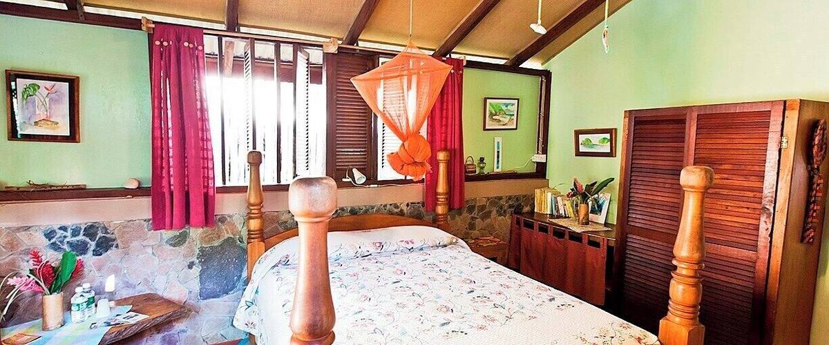 A wooden bed with a floral bedspread, a woven mat, a desk with a plant, and a wooden wardrobe.