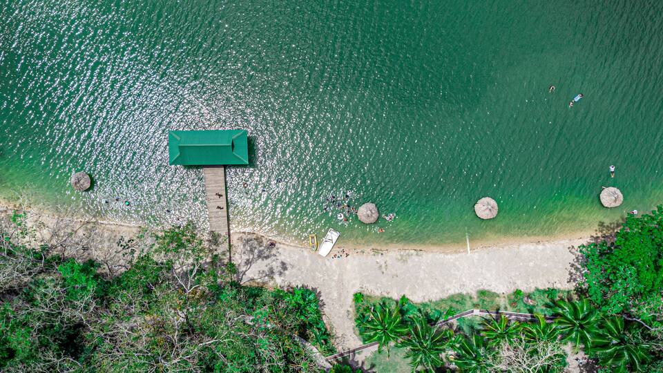 An aerial view of a beach with a wooden pier, clear turquoise water, and palm trees.