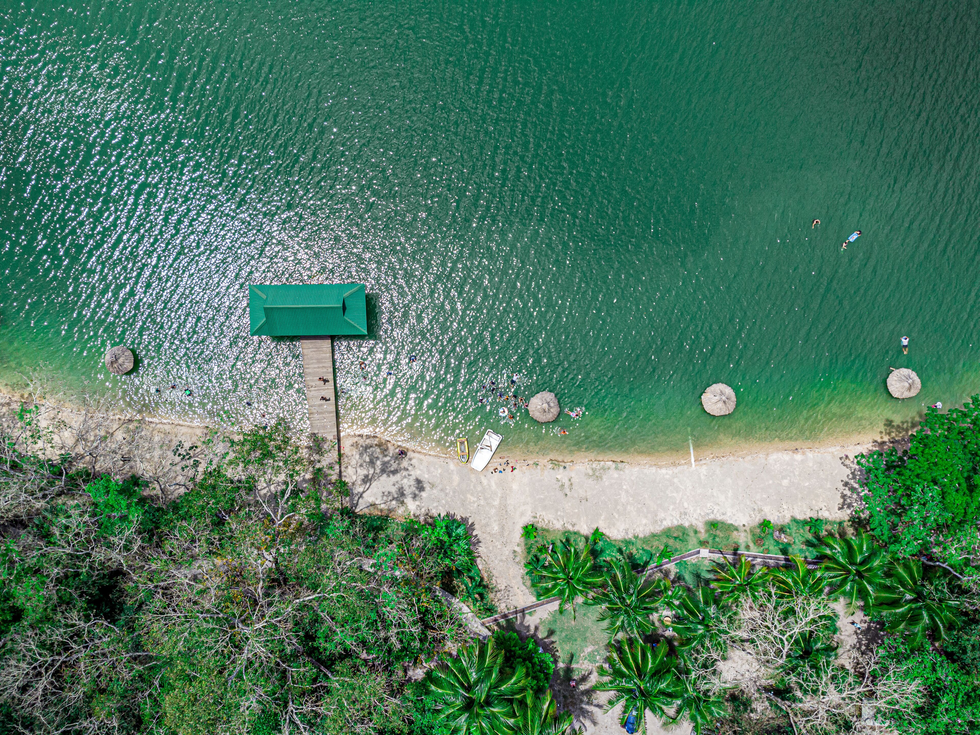 An aerial view of a beach with a wooden pier, clear turquoise water, and palm trees.
