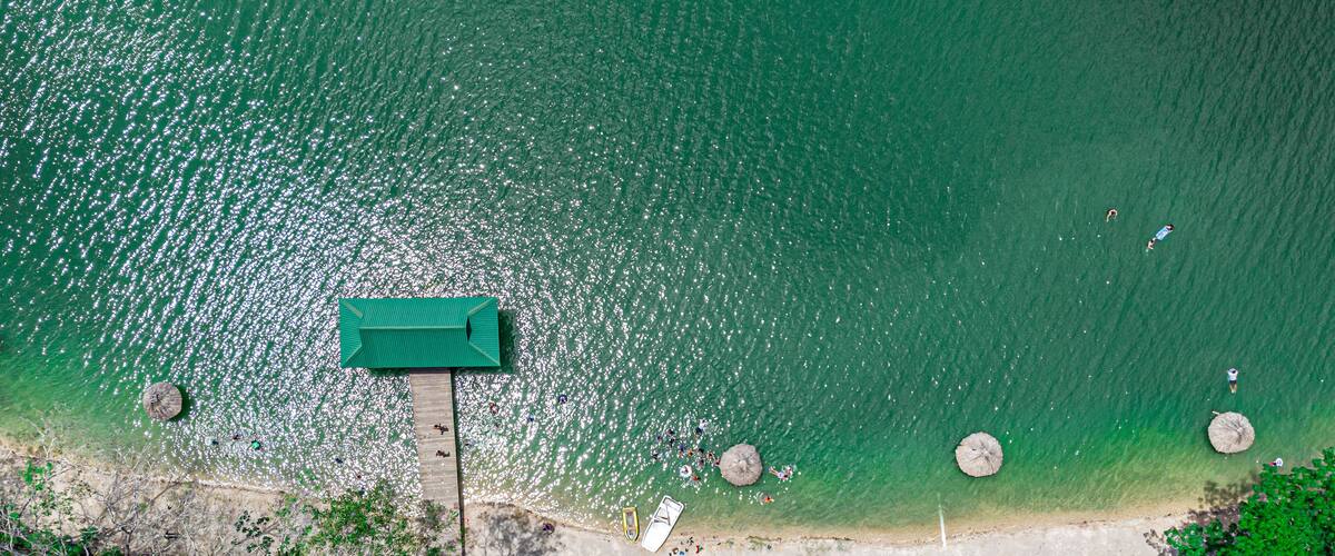 An aerial view of a beach with a wooden pier, clear turquoise water, and palm trees.