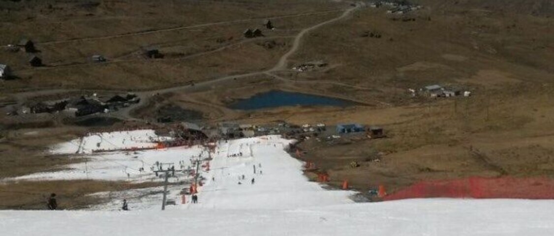 Un paisaje nevado con una pista de esquí, un lago azul pequeño y algunos edificios a lo lejos.