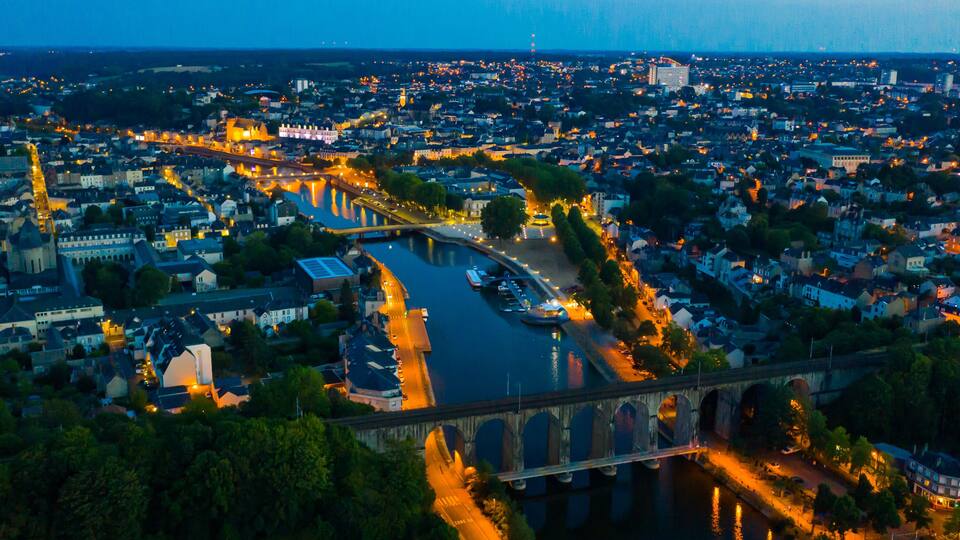 An aerial view of a city at dusk with a river, a bridge, and illuminated streets.