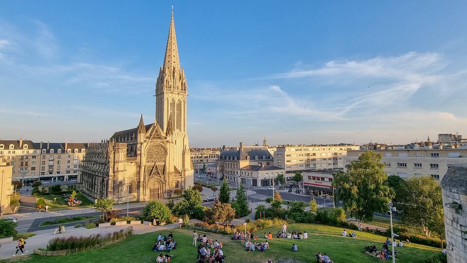 Una catedral con una aguja alta, rodeada por un paisaje urbano con edificios y un parque con gente.