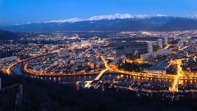 A cityscape at dusk with a river, illuminated buildings, and snow-capped mountains in the background.