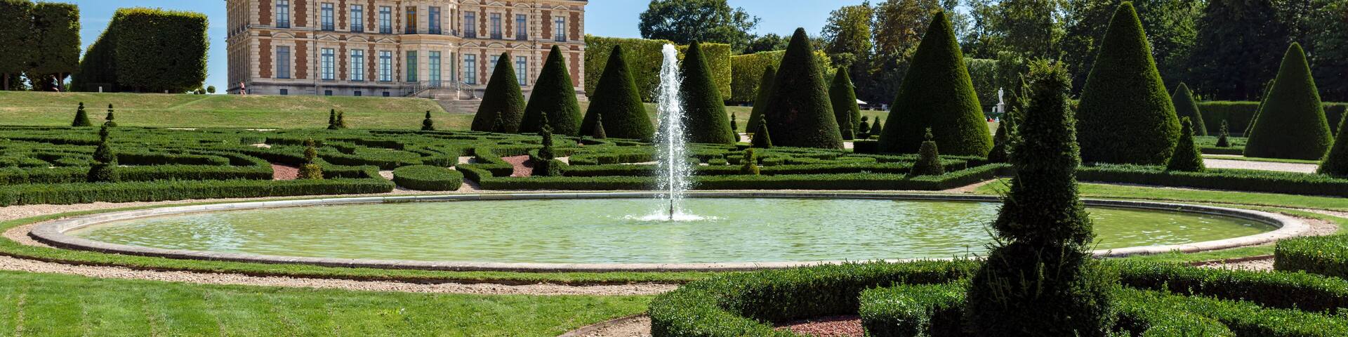 A grand château with a formal garden, including a fountain and geometrically trimmed hedges.