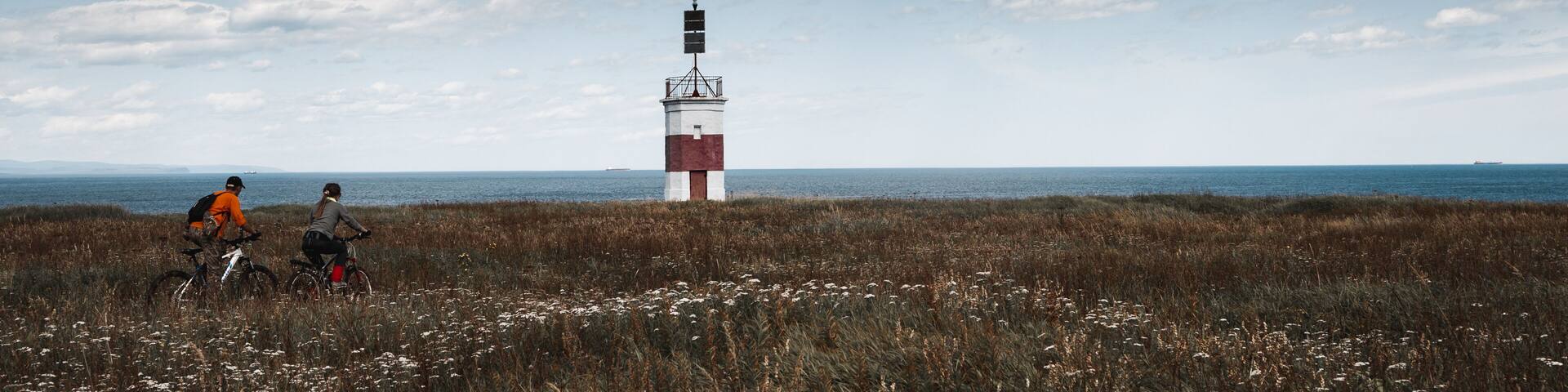 A coastal landscape with a lighthouse and two cyclists in the foreground.
