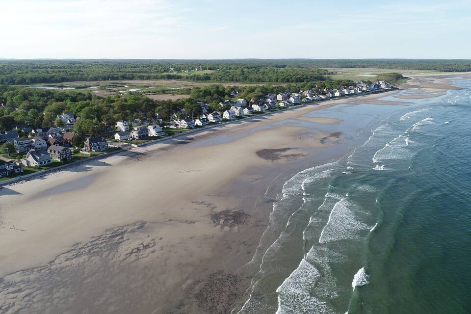 An aerial view of a coastal town with a long sandy beach, houses along the shore, and a forested area in the background.