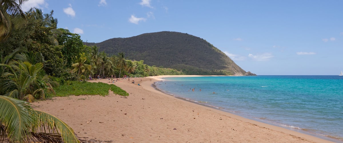 A beach with clear turquoise water, a sandy shore, and a mountain in the background.