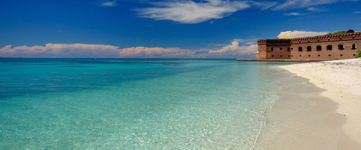 A beach with clear turquoise water and a large brick building on the shore.