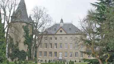 Um edifício grande e histórico com fachada simétrica, uma torre do relógio central e um jardim formal com sebes bem aparadas e um caminho de cascalho.