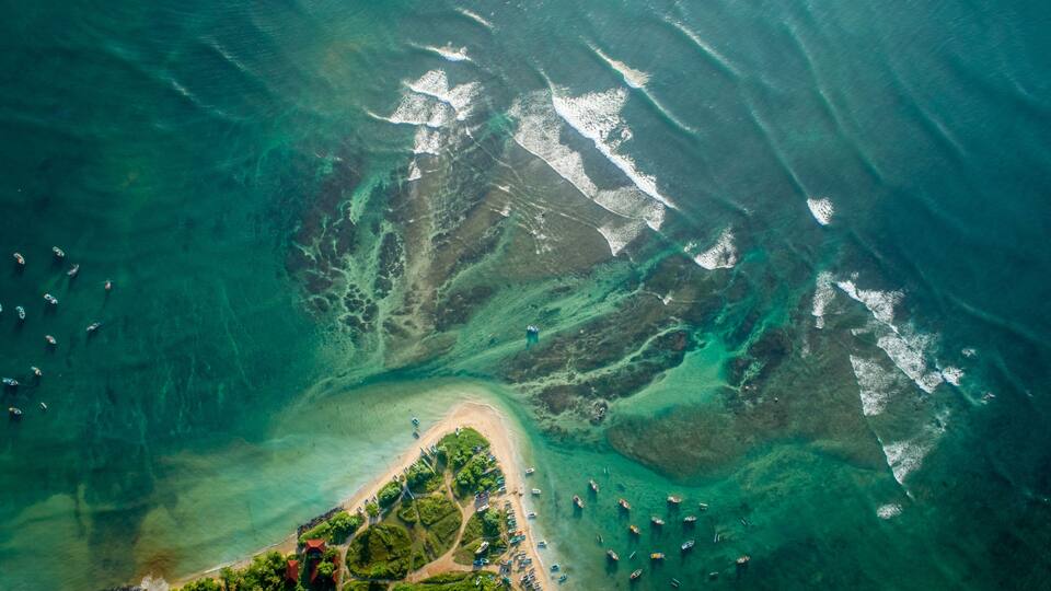 An aerial view of a coastal area with clear turquoise waters, a sandy beach, and numerous boats.