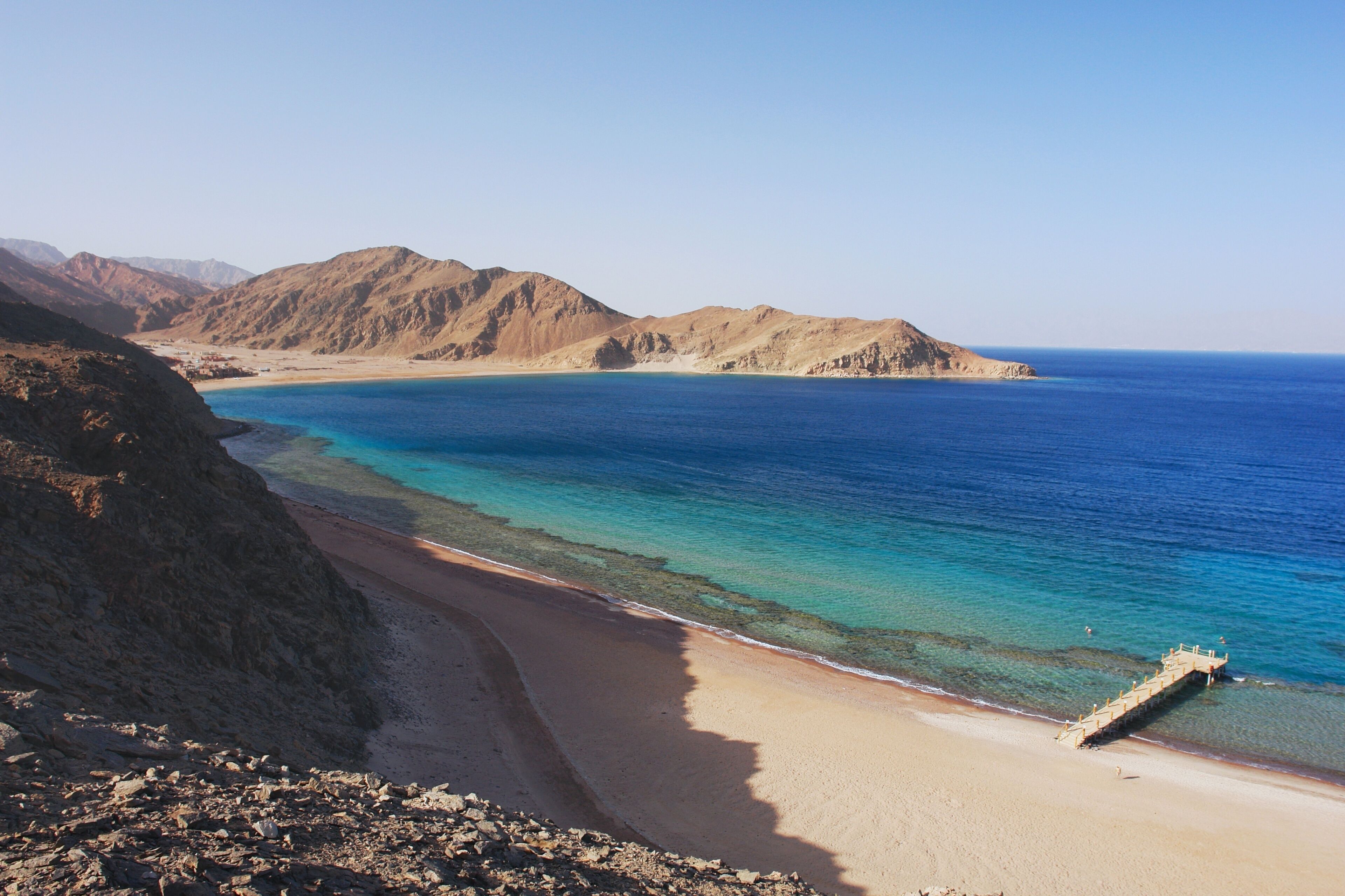 A coastal landscape with a sandy beach, clear blue waters, and rocky hills in the background.