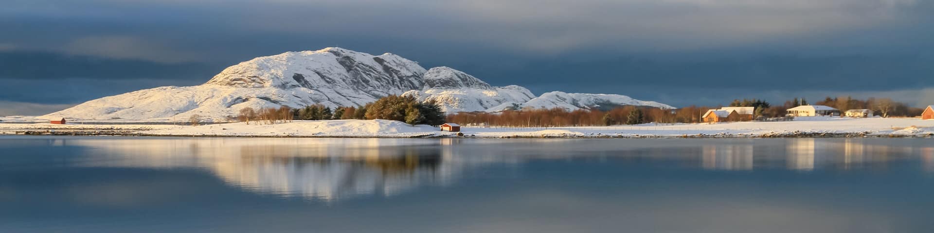 A snow-covered mountain, a calm lake, and a small village with buildings.