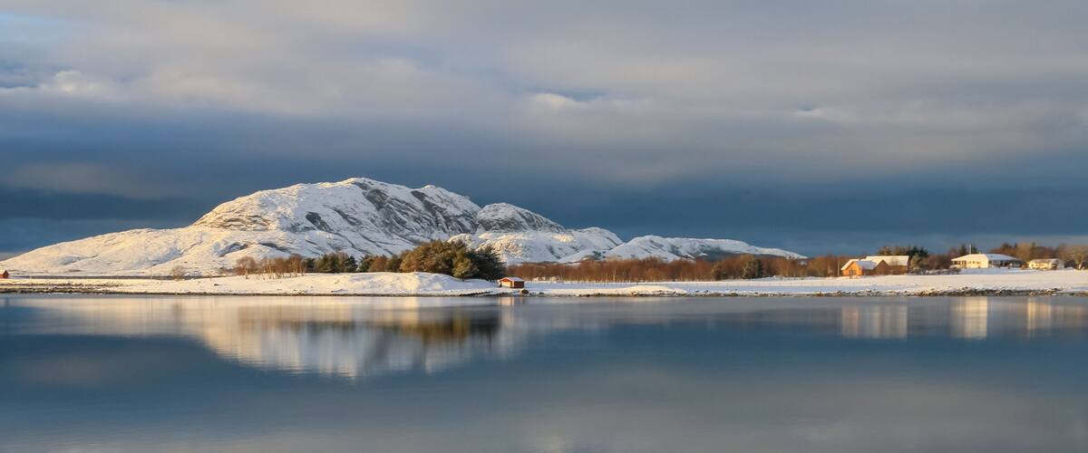 A snow-covered mountain, a calm lake, and a small village with buildings.
