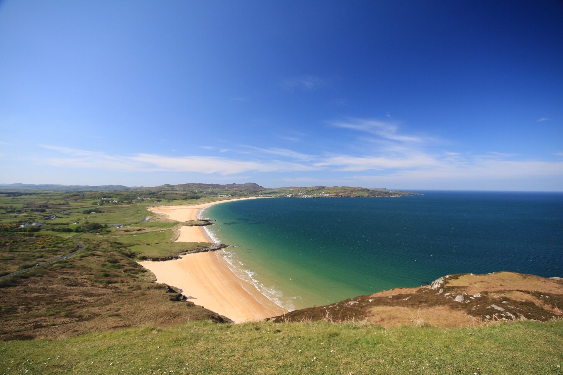A coastal landscape with a sandy beach, green hills, and a clear blue sky.