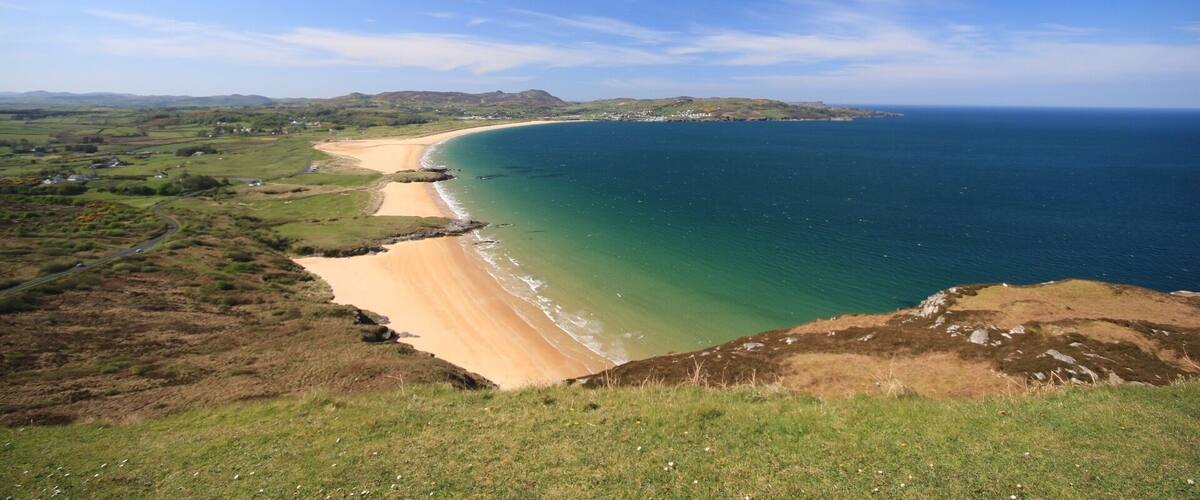 A coastal landscape with a sandy beach, green hills, and a clear blue sky.