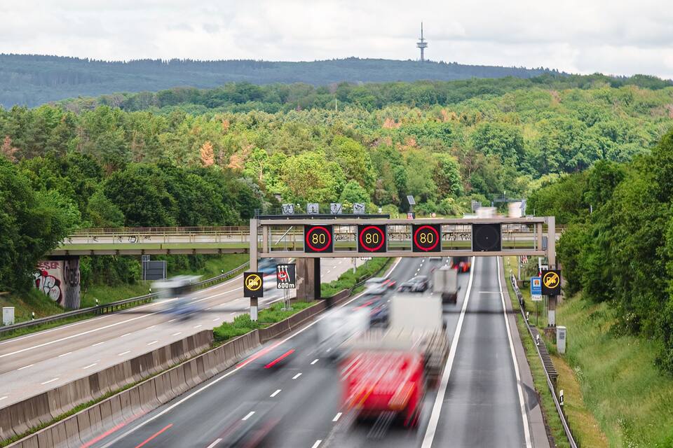 Una autopista con señales de límite de velocidad, un puente y una ladera boscosa al fondo.
