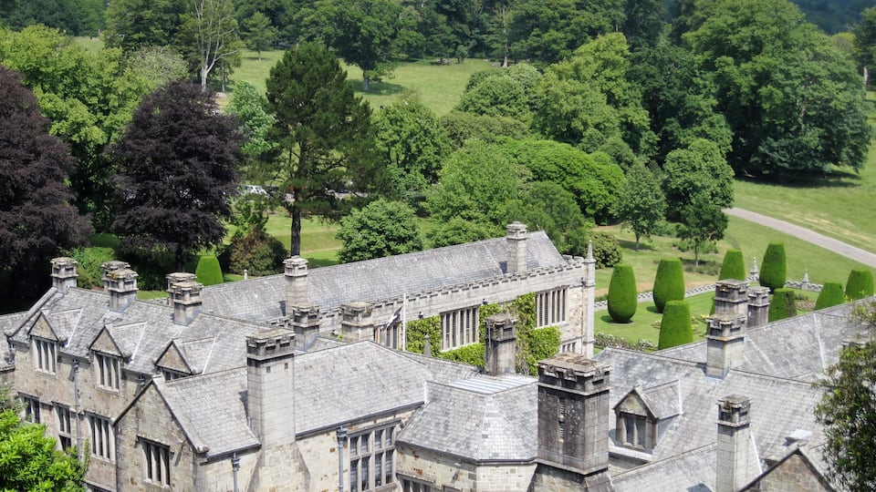 A historic stone building with a complex roof and multiple chimneys, surrounded by lush greenery and a well-maintained lawn.